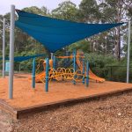Playground structure under bright blue shade set in bark mulch with bright orange slides