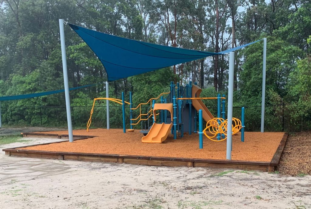 Playground structure under bright blue shade set in bark mulch with bright orange slides
