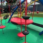 Multiple hanging climbers with red pods on black chain and a large red plastic bucket swing