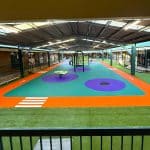 Undercover play area with bright orange, purple and turquoise rubber surfacing surrounded by synthetic grass