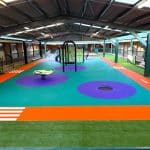 Undercover play area with bright orange, purple and turquoise rubber surfacing surrounded by synthetic grass
