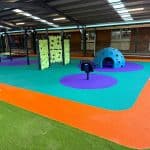 Undercover play area with bright orange, purple and turquoise rubber surfacing surrounded by synthetic grass