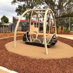 Inclusive playground swing with wheelchair access, set on soft surfacing and framed by curved metal bars. Surrounded by wood chips, paved paths, trees, and nearby buildings, the space supports accessible outdoor play.