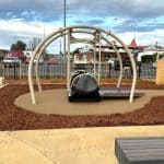 Inclusive playground swing with wheelchair access, set on soft surfacing and framed by curved metal bars. Surrounded by wood chips, paved paths, trees, and nearby buildings, the space supports accessible outdoor play.