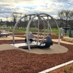 Inclusive playground swing with wheelchair access, set on soft surfacing and framed by curved metal bars. Surrounded by wood chips, paved paths, trees, and nearby buildings, the space supports accessible outdoor play.