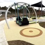 Inclusive playground featuring a modern swing set with one accessible seat designed for children with disabilities, supported by a curved metal frame. The play area has a soft, rubberised yellow surface with circular patterns for safety and visual appeal. The playground promotes inclusive outdoor play in a thoughtfully designed community space.