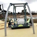 Inclusive playground featuring a modern swing set with one accessible seat designed for children with disabilities, supported by a curved metal frame. The play area has a soft, rubberised yellow surface with circular patterns for safety and visual appeal. In the background, wooden climbing structures and shaded canopies provide additional recreational options. Surrounded by grassy areas, a paved path, and nearby residential buildings, the playground promotes inclusive outdoor play in a thoughtfully designed community space.
