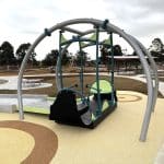 Inclusive playground featuring a modern swing set with one accessible seat designed for children with disabilities, supported by a curved metal frame. The play area has a soft, rubberised yellow surface with circular patterns for safety and visual appeal. In the background, wooden climbing structures and shaded canopies provide additional recreational options. Surrounded by grassy areas, a paved path, and nearby residential buildings, the playground promotes inclusive outdoor play in a thoughtfully designed community space.