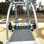 Inclusive playground featuring a modern swing set with one accessible seat designed for children with disabilities, supported by a curved metal frame. The play area has a soft, rubberised yellow surface with circular patterns for safety and visual appeal. The playground promotes inclusive outdoor play in a thoughtfully designed community space.