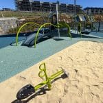 Modern residential playground featuring a sandy area with a green and black spring rider, rubberised safety surface, and a large climbing structure with slides, nets, and curved metal bars. Surrounded by fencing and set against a backdrop of new residential buildings and construction under clear daylight
