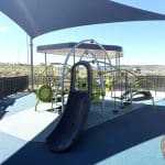 Contemporary playground with a blue rubber safety surface, featuring a dark blue slide, climbing ropes, curved metal bars, and green interactive panels. A large shade canopy provides sun protection, and the area is enclosed by a black metal fence with open landscape and distant buildings in the background.
