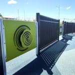 Outdoor playground area with blue rubber flooring, interactive sensory panels, and black metal fencing casting long shadows in bright sunlight. Urban buildings and light poles visible in the background.