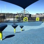 Modern playground with blue rubber safety flooring in varied patterns, featuring three green and blue stepping play elements in the foreground. A large climbing frame with netting and a slide sits behind a fenced area, shaded by an overhead canopy. Surrounding the playground are buildings and a clear blue sky.