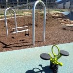 Playground area with a green and black two-seat spring rider in the foreground, alongside a swing set with a flat seat and toddler bucket seat. The surface combines rubber safety flooring and mulch, with stone steps and young landscaping leading up a small hill. Construction scaffolding and buildings appear in the background under a clear sky.