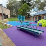 Outdoor play area with purple and blue rubber wetpour surfacing featuring multiple play options for inclusivity. roller table, cozy dome in a bright green colour, rubber domes, spinning chair and climbing wall