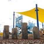 Outdoor playground with concrete wooden look stepping stumps, blue climbing structures, slides, and yellow shade sails over a bark mulch surface