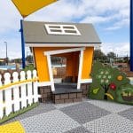 Bright orange toddler cubby house with white picket fence and green bush with bells