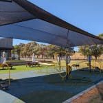 Outdoor gym area with yellow and black exercise equipment including squat press, ab crunch and mobility devices set on a blue and green rubber safety surface. A large shade structure covers the gym, with picnic tables, umbrellas, and a modern concrete building in the background. Surrounded by open fields and trees under a clear blue sky