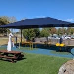 Outdoor gym area with yellow and black exercise equipment including squat press, ab crunch and mobility devices set on a blue and green rubber safety surface. A large shade structure covers the gym, with picnic tables, umbrellas, and a modern concrete building in the background. Surrounded by open fields and trees under a clear blue sky