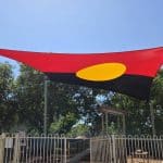 Hypar Sail shade canopy inspired by the Aboriginal flag, featuring red and black halves with a central yellow sun circle, installed above a fenced playground in a community pre school in Australia, symbolizing Indigenous culture and inclusion.