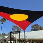 Hypar Sail shade canopy inspired by the Aboriginal flag, featuring red and black halves with a central yellow sun circle, installed above a fenced playground in a community pre school in Australia, symbolizing Indigenous culture and inclusion.