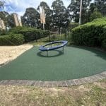 Green Rubber Wetpour surfacing surrounding a childrens outdoor ring spinning play item with a screening hedge to the sides under clear skies