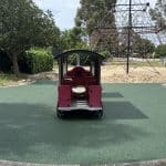 Green Rubber Wetpour surfacing surrounding a childrens outdoor red train play item with a pyramid net climber in sand in the background under clear skies