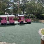 Green Rubber Wetpour surfacing surrounding a childrens outdoor red train play item with tall trees in the background under clear skies