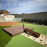 Outdoor water play area featuring a manual hand pump, timber platform, and stone-lined trough—encouraging sensory exploration and STEM learning in a safe, inclusive playground with artificial turf and secure fencing.