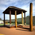 Nature-inspired playground structure with timber posts, thatched roof pavilion, and suspended rope bridge—encouraging imaginative play and physical development in a safe, outdoor setting with soft-fall surfacing.