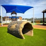 Nature-themed playground featuring a hollow log tunnel on artificial turf, with shaded climbing structures and soft-fall surfacing—encouraging imaginative exploration and safe outdoor play in a well-maintained early learning environment.