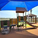 Children’s playground with climbing structure, slide, bridge walkway, and interactive play panel under a large blue shade canopy—supporting physical activity, imaginative play, and safe outdoor learning in a well-maintained recreational space.