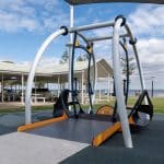 Wheelchair-accessible playground swing with ramp access in a public park, promoting inclusive play for people with mobility impairments. Features sturdy metal frame in grey, blue, and orange, with picnic tables and water views in the background