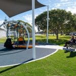 Person pushing a wheelchair user along a paved path toward an inclusive playground swing with ramp access, under a shade structure in a green park setting. Highlights accessible design and community engagement in outdoor recreational spaces