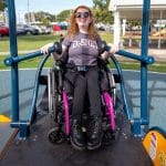 Smiling individual in a wheelchair using inclusive playground equipment with textured platform and handrails, wearing a Disney Club t-shirt. Set in a park with pavilion, trees, and parked cars, showcasing accessible recreational design