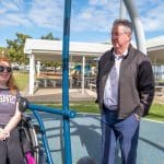 Three people interacting at an accessible playground with rubberised surface and shade canopy. One person in a wheelchair wearing a Disney Club t-shirt, engaging with two standing individuals. Emphasises inclusive community spaces and social connection