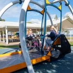 Two individuals on a dual-seat inclusive playground swing—one in a wheelchair and one on a standard seat—supported by curved white and blue metal frame. Located in a waterfront park with pavilion and trees, promoting accessible play for all abilities.