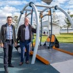 Two people beside a large wheelchair-accessible playground swing with ramp set on a rubberised surface in a waterfront park. Surrounded by grass, trees, and shaded seating, highlighting inclusive design for people with mobility challenges.