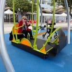 Woman enjoying an accessible wheelchair swing in an inclusive public playground with safety surfacing, vibrant green and orange accents, and modern urban surroundings—highlighting disability-friendly recreational equipment and inclusive community design