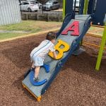 Play equipment with a single deck and a double navy blue slide, navy blue roof and multiple climbers including one with the letters A, B, C on the ramp in bright primary colours and a toddler climbing up