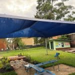 A shaded outdoor learning space featuring a water play station with a hand‑crank, a dry creek bed with rocks, a small wooden bridge, and landscaped garden beds. The area includes educational displays, a green shed, and natural play elements designed for exploration in a school or childcare environment.