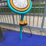 a musical drum for outdoor use set in rubber surfacing it has a blue frame with a small circular head with a steel drum face and mallet hanging on the holder attached to a single support post.