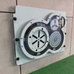 An educational wall‑mounted gear panel featuring interlocking mechanical gears behind a clear protective cover. Installed above green rubber surfacing, this STEM‑focused play element encourages hands‑on learning and fine‑motor development in school, childcare, and community play environments.
