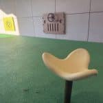 A children’s play area with green rubber safety surfacing featuring a beige spinning seat and two wall‑mounted activity panels: a vehicle dashboard with steering wheel and gauges, and a green maze puzzle. Designed to promote imaginative, sensory, and fine‑motor play in indoor or covered outdoor environments.