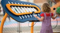 Small girl in pink dress holding striker in her left hand standing in front of a large blue xylophone board playing music
