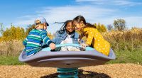 3 children sitting on large circular purple dish with blue handles holding on while it bounces